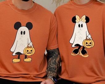 May include: Two orange t-shirts featuring a cartoon ghost character with Mickey Mouse ears and a pumpkin bucket. The ghost is white with black eyes and feet. One ghost has a bow. The shirts are short-sleeved.