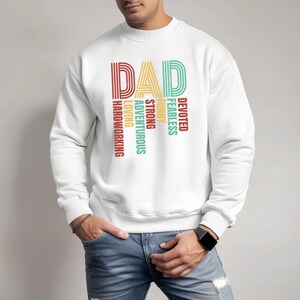May include: White sweatshirt with the word "DAD" in large, colorful letters. The letters are made up of smaller words describing a father, such as "devoted", "fearless", "funny", "strong", "adventurous", "loving", and "hardworking".