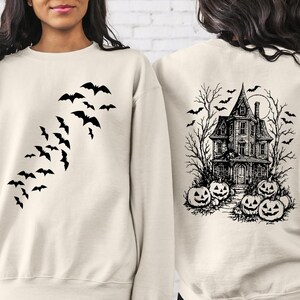 May include: A cream-colored crewneck sweatshirt featuring a black graphic print. The front displays a flock of flying bats. The back showcases a detailed black ink illustration of a haunted house surrounded by bare trees, flying bats, and jack-o'-lanterns.