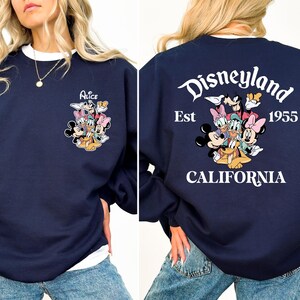May include: Navy blue sweatshirt with white trim featuring a Disney character graphic on the front and back. The front graphic says "Alice" with Disney characters. The back graphic says "Disneyland Est 1955 California" with Disney characters.