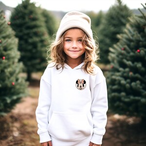K&ouml;nnte beinhalten: Wei&szlig;er Kapuzenpullover mit einem Minnie-Maus-Motiv auf der Brust. Das Motiv enth&auml;lt eine Schleife und einen karierten Hintergrund. Der Pullover hat eine Vordertasche und wird mit einer cremefarbenen M&uuml;tze getragen.