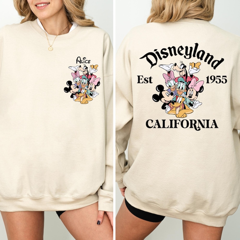 May include: Cream-colored sweatshirt featuring a Disney character graphic on the front and back. The front displays a group of characters with the name "Alice" above them. The back has the Disneyland logo, "Est 1955" and "California".