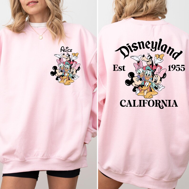 May include: A light pink sweatshirt featuring a Disney character graphic on the front and back. The front graphic includes the name "Alice" above a group of characters. The back graphic says "Disneyland Est 1955 California".