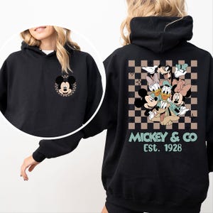K&ouml;nnte beinhalten: Schwarzer Kapuzenpullover mit einem Mickey Mouse-Motiv auf der Vorderseite und einer Gruppe von Disney-Figuren auf der R&uuml;ckseite. Das R&uuml;ckendesign ist vor einem braunen und beigefarbenen Karomuster platziert, mit dem Aufdruck "Mickey & Co Est. 1928".