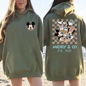 K&ouml;nnte beinhalten: Olivgr&uuml;ner Kapuzenpullover mit Mickey Mouse auf der Vorderseite und einer Gruppe von Disney-Figuren auf der R&uuml;ckseite. Das R&uuml;ckendesign zeigt Mickey, Minnie, Donald, Daisy und Goofy vor einem karierten Hintergrund. Der Text "Mickey & Co Est. 1928" ist darunter gedruckt.