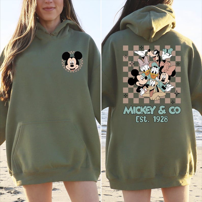 K&ouml;nnte beinhalten: Olivgr&uuml;ner Kapuzenpullover mit Mickey Mouse auf der Vorderseite und einer Gruppe von Disney-Figuren auf der R&uuml;ckseite. Das R&uuml;ckendesign zeigt Mickey, Minnie, Donald, Daisy und Goofy vor einem karierten Hintergrund. Der Text "Mickey & Co Est. 1928" ist darunter gedruckt.