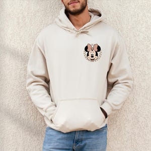 K&ouml;nnte beinhalten: Beige Kapuzenpullover mit einer Vordertasche und einer Kapuze mit Kordelzug. Der Kapuzenpullover hat eine Minnie Mouse Grafik auf der oberen linken Brust. Die Grafik ist ein kreisf&ouml;rmiges Design mit kariertem Hintergrund und einem Minnie Mouse Gesicht.