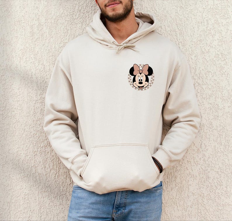 K&ouml;nnte beinhalten: Beige Kapuzenpullover mit einer Vordertasche und einer Kapuze mit Kordelzug. Der Kapuzenpullover hat eine Minnie Mouse Grafik auf der oberen linken Brust. Die Grafik ist ein kreisf&ouml;rmiges Design mit kariertem Hintergrund und einem Minnie Mouse Gesicht.
