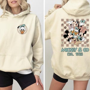 K&ouml;nnte beinhalten: Cremefarbener Kapuzenpullover mit Fronttasche und Kordelzugkapuze. Auf der Vorderseite befindet sich eine Donald Duck-Grafik in einem karierten Kreis. Auf der R&uuml;ckseite ist eine Gruppe von Disney-Figuren mit dem Text "Mickey & Co. Est. 1928" in T&uuml;rkis abgebildet.