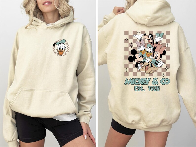 K&ouml;nnte beinhalten: Cremefarbener Kapuzenpullover mit Fronttasche und Kordelzugkapuze. Auf der Vorderseite befindet sich eine Donald Duck-Grafik in einem karierten Kreis. Auf der R&uuml;ckseite ist eine Gruppe von Disney-Figuren mit dem Text "Mickey & Co. Est. 1928" in T&uuml;rkis abgebildet.