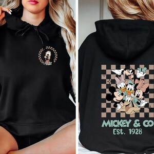 K&ouml;nnte beinhalten: Schwarzer Kapuzenpullover mit einem kleinen Goofy-Motiv auf der Vorderseite. Auf der R&uuml;ckseite befindet sich eine gr&ouml;&szlig;ere Grafik von Mickey, Minnie, Donald, Daisy und Goofy mit dem Text "Mickey & Co Est. 1928". Das Design ist auf einem karierten Hintergrund.