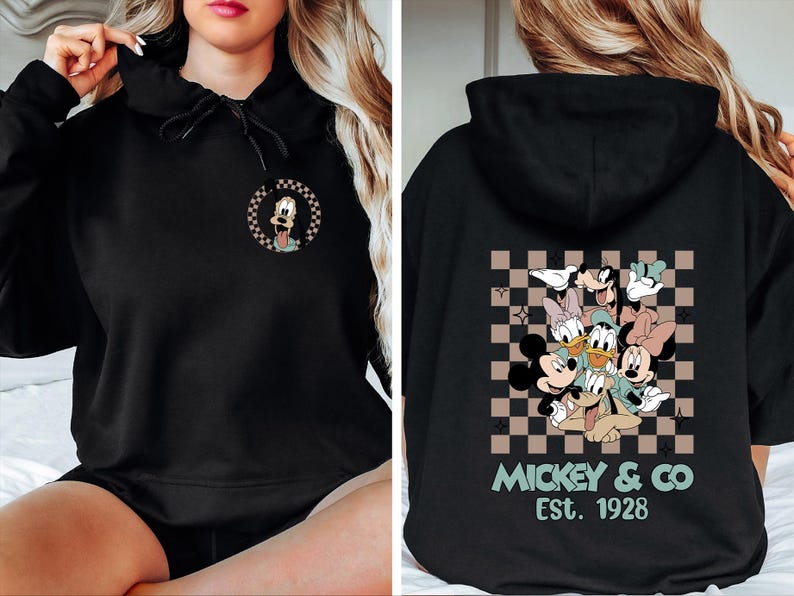 K&ouml;nnte beinhalten: Schwarzer Kapuzenpullover mit einem kleinen Goofy-Motiv auf der Vorderseite. Auf der R&uuml;ckseite befindet sich eine gr&ouml;&szlig;ere Grafik von Mickey, Minnie, Donald, Daisy und Goofy mit dem Text "Mickey & Co Est. 1928". Das Design ist auf einem karierten Hintergrund.