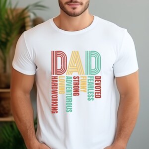 May include: White t-shirt with the word "DAD" in colorful retro-style lettering. The letters are made up of vertical stripes. Below the word "DAD" are the words "Devoted, Fearless, Funny, Strong, Adventurous, Loving, Hardworking" in a vertical column.