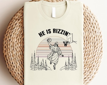 May include: A beige t-shirt with a black and white illustration of a person playing basketball. The text "HE IS RIZZIN'" is printed on the shirt.