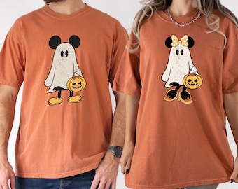 May include: Two orange t-shirts featuring a cartoon ghost with Mickey Mouse ears holding a jack-o'-lantern. One ghost has a bow. The shirts are short-sleeved and have a vintage, distressed look, perfect for Halloween.