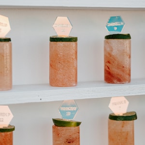 Op de afbeelding: Een display van decoratieve zout shotglazen, elk bekroond met een limoenschijfje en een zeshoekig naambordje. De shotglazen zijn gerangschikt op witte planken. De naambordjes hebben namen en nummers.