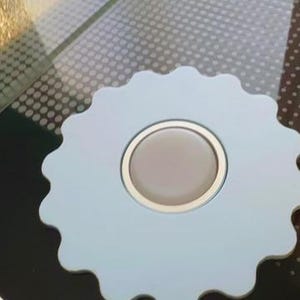 May include: A light blue, scalloped-edge coaster with a circular, beige center and a gold-colored rim. The coaster is set on a dark surface, with a patterned background visible in the upper right corner.