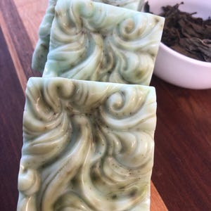 May include: Four bars of green and white swirl soap with a decorative design. The soap is on a wooden surface.