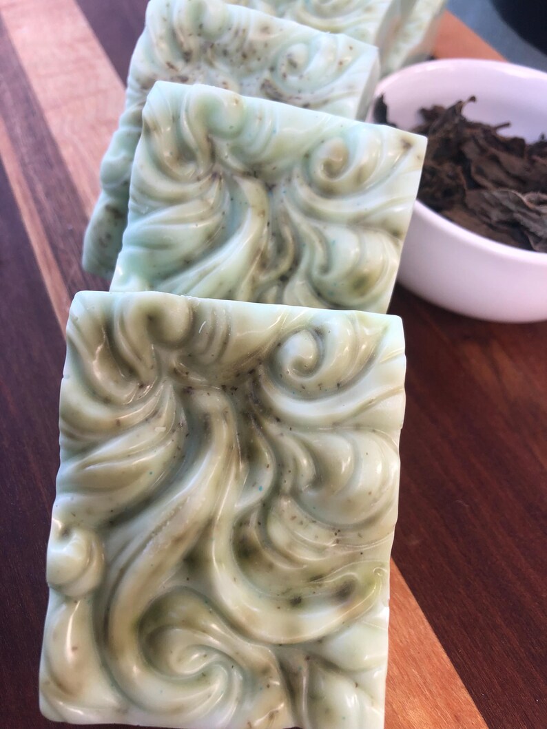 May include: Four bars of green and white swirl soap with a decorative design. The soap is on a wooden surface.