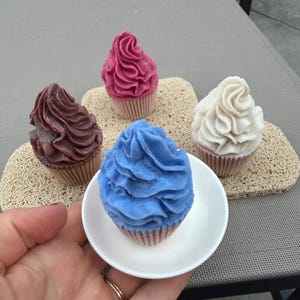 May include: Four cupcake-shaped soaps with swirled frosting tops in shades of dark purple, magenta, bright blue, and white. Each soap sits in a pleated paper-style liner. The blue soap is held in a small white dish, while the others rest nearby.