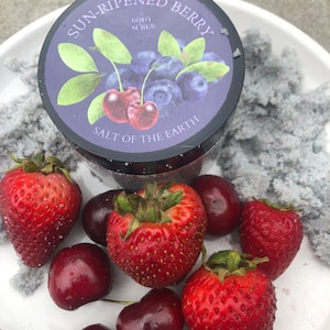 May include: A jar of body scrub with a purple label that says "Sun-Ripened Berry Body Scrub Salt of the Earth". The label has a drawing of blueberries and cherries. The jar is surrounded by red strawberries and cherries on a white surface.