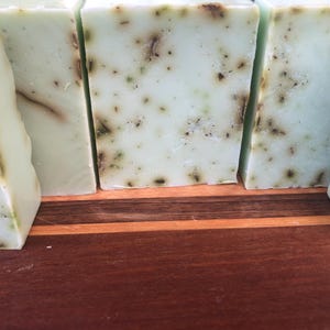 May include: Four bars of handmade soap with a light green color and flecks of green throughout. The soap bars are sitting on a wooden surface.