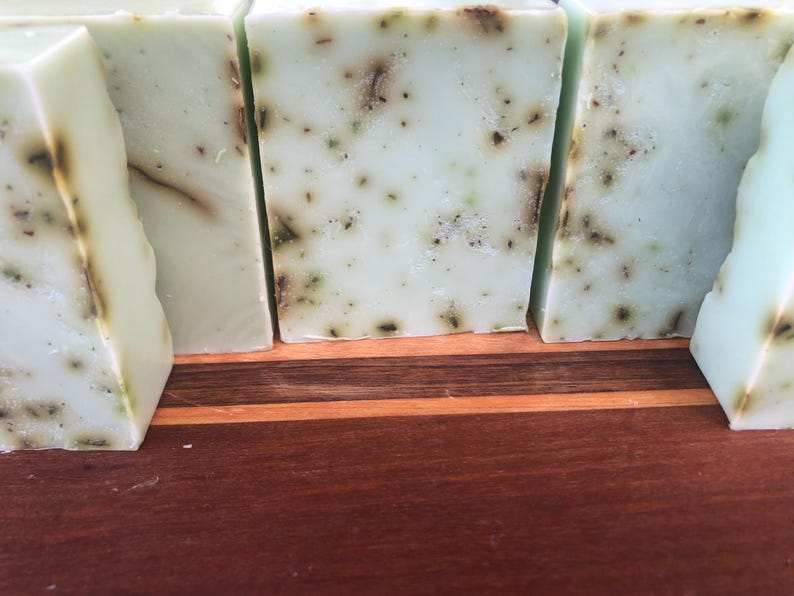 May include: Four bars of handmade soap with a light green color and flecks of green throughout. The soap bars are sitting on a wooden surface.