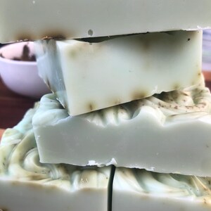 May include: Four bars of handmade soap, stacked on top of each other. The soap is a light green color with a swirl pattern and flecks of brown throughout.