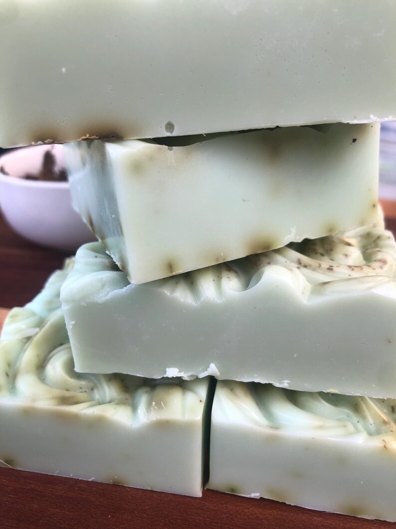 May include: Four bars of handmade soap, stacked on top of each other. The soap is a light green color with a swirl pattern and flecks of brown throughout.