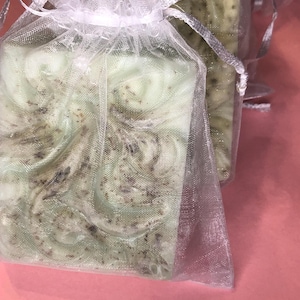 May include: Two bars of green soap with flecks of brown, wrapped in a white mesh bag. The soap is sitting on a pink surface.
