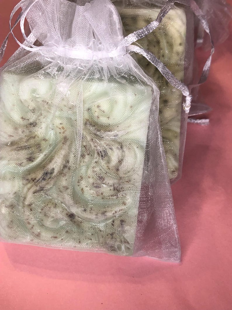 May include: Two bars of green soap with flecks of brown, wrapped in a white mesh bag. The soap is sitting on a pink surface.