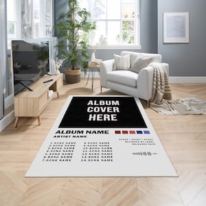 May include: A white rug with a black rectangle in the center that says "ALBUM COVER HERE" in white text. Below the rectangle is a black and white list of song titles and an artist name. The rug is on a wooden floor.