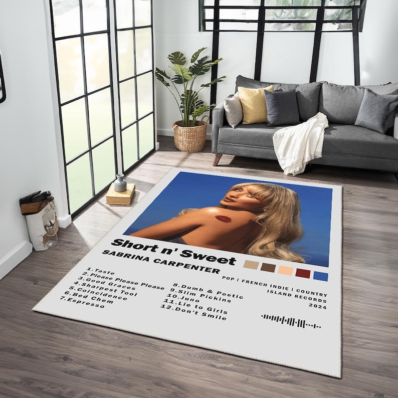 May include: A white rug with a blue background featuring a woman with blonde hair. The rug has a black and white graphic with the text "Short n' Sweet" and "Sabrina Carpenter". The graphic also includes a list of song titles and the album's release information.