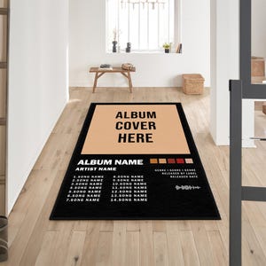 Puede incluir: Alfombra rectangular negra con un diseño de portada de álbum en color beige y el texto "ALBUM COVER HERE". Incluye "ALBUM NAME" y "ARTIST NAME", además de títulos de canciones y una sección para género, sello y fecha de lanzamiento.