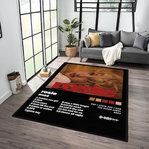 May include: Black rectangular area rug featuring a portrait of a woman with blonde hair and the name "rosie" in red. The rug includes a tracklist and album details. The rug is placed in a living room setting.