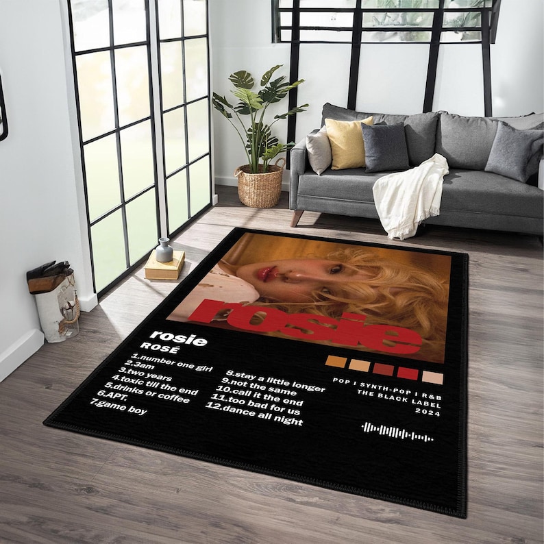May include: Black rectangular area rug featuring a portrait of a woman with blonde hair and the name "rosie" in red. The rug includes a tracklist and album details. The rug is placed in a living room setting.