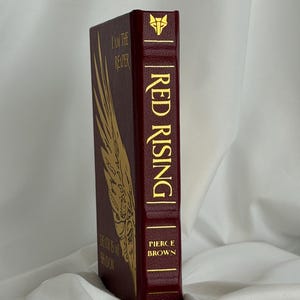 Peut inclure: Un livre relié intitulé Red Rising de Pierce Brown avec une couverture rouge foncé et des lettres dorées. Le dos présente un emblème de loup, le titre, le nom de l'auteur et le chiffre un. La couverture affiche un grand motif d'aile dorée.
