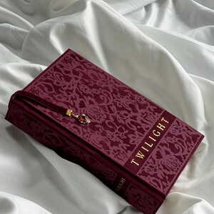 May include: A hardcover book titled Twilight with a deep burgundy velvet cover featuring an intricate floral pattern. The book includes a matching burgundy ribbon bookmark with a small gold-toned charm containing a red stone accent.