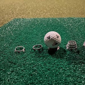 May include: A set of four golf tee holders in various sizes, with a white golf ball on top of the largest holder. The holders are made of a white plastic material with a textured surface. The golf ball has a white and black design.
