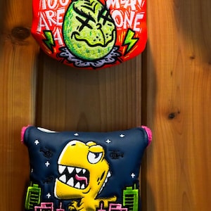 May include: Two golf club head covers. The top cover is orange with a green cartoon character wearing a Santa hat and the text "You Are A Mean One". The bottom cover is dark blue with a yellow cartoon dinosaur in front of a city skyline.