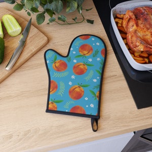 May include: A blue oven mitt with a peach pattern. The mitt features a repeating design of ripe peaches, green leaves, and small white flowers. The mitt has a black trim and a loop for hanging.