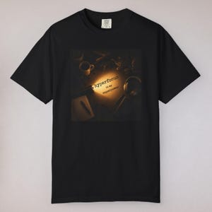 Może przedstawiać: Czarny t-shirt z krótkim rękawem z centralną grafiką przedstawiającą scenę na biurku oświetloną jasnym reflektorem. Grafika zawiera kubek, słuchawki i długopis, z napisem "hyperfocus is my superpower" wydrukowanym w świetle.