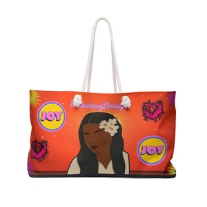May include: A large orange tote bag with white rope handles featuring a central illustration of a person with dark hair and white flowers. The bag displays the text TreasuresofBeauty, two yellow circles with the word JOY, and two black hearts.