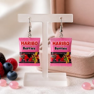 Può includere: Un paio di orecchini pendenti con ciondoli in miniatura a forma di sacchetto di caramelle Haribo Berries rosa. Ogni ciondolo mostra il logo Haribo Berries in rosso e nero, insieme a un orsetto giallo e diverse bacche disegnate.