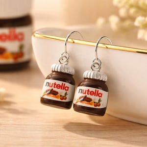 Peut inclure: Boucles d'oreilles pendantes en forme de petits pots de Nutella. Les boucles d'oreilles ont des crochets argentés et sont marron avec des étiquettes blanches qui indiquent "nutella". Un pot de Nutella et des fleurs blanches sont en arrière-plan.