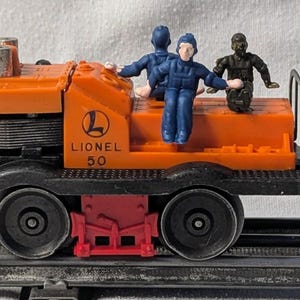 May include: An orange Lionel 50 toy train engine with black wheels and a red undercarriage. The train has three small figurines on top. The train is on a gray track.