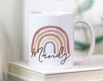 May include: White ceramic mug featuring a watercolor rainbow design in shades of brown, tan, and green. The name "Mandy" is written in a cursive font below the rainbow. The mug is sitting on a stack of books.