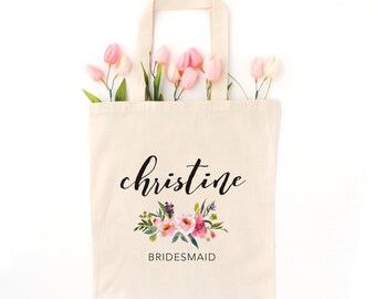 Puede incluir: Bolso tote de color crema con el nombre "christine" en escritura negra y un diseño floral. La palabra "BRIDESMAID" está debajo del diseño floral. Hay tulipanes rosas dentro de la bolsa.