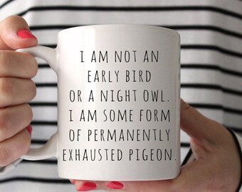 Può includere: Tazza in ceramica bianca con testo nero: "I AM NOT AN EARLY BIRD OR A NIGHT OWL. I AM SOME FORM OF PERMANENTLY EXHAUSTED PIGEON." La tazza è tenuta da una persona con lo smalto rosso.