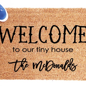 May include: A rectangular doormat made of natural fibers, with the words "WELCOME to our tiny house the McDonalds" in black script. Blue canvas shoes with white soles are placed on the top left corner.